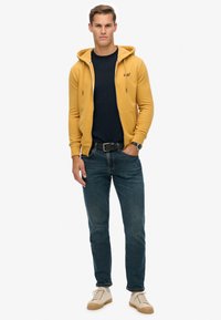 Giovane uomo in piedi che indossa una felpa con zip gialla senape, t-shirt nera, jeans blu, cintura nera, orologio e sneakers beige.