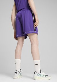 Pantalones cortos deportivos de malla morados con ribetes blancos, que presentan rayas laterales. Se llevan con calcetines blancos y unas zapatillas elegantes y multicolores.
