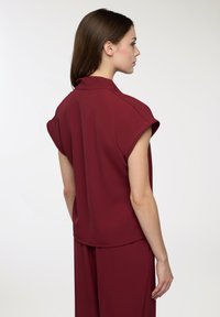 Blusa corta bordeaux a maniche corte in tessuto morbido, con colletto e vestibilità rilassata. Dettaglio notevole include cuciture sottili lungo la spalla.