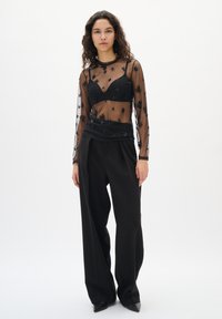 Frau mit lockigem Haar, die ein durchsichtigen schwarzen Langarmtop mit floralem Stickerei, einen schwarzen Bralette, weit geschnittene schwarze Hose und schwarze spitze Schuhe trägt.