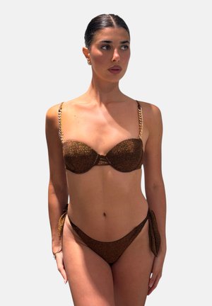 Donna con capelli scuri in bikini marrone glitterata con spalline con catena dorata, che guarda a sinistra su uno sfondo chiaro uniforme.
