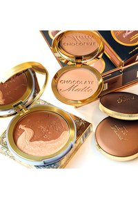 Twee compacte bronzers in gouden doosjes, de ene met een pauwenontwerp en bloemmotieven, in matte bruin- en karamelkleuren, naast een doos.