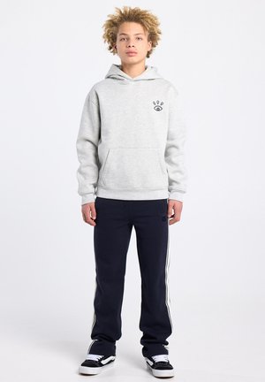 Grijze hoodie met een zak aan de voorkant en een klein grafisch ontwerp op de borst, gecombineerd met marineblauwe joggingbroek met witte strepen aan de zijkant en zwarte sneakers.