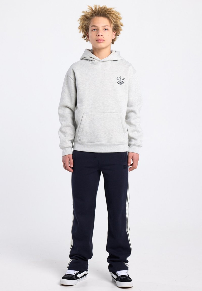 Grauer Kapuzenpullover mit einer Vordertasche und einer kleinen Grafik auf der Brust, kombiniert mit marineblauen Jogginghosen mit weißen Seitenstreifen und schwarzen Turnschuhen.