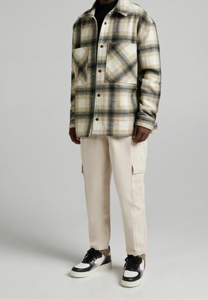 Mężczyzna w beżowej, kraciastej zapinanej na guziki kurtce, jasnobeżowych spodniach typu cargo i czarno-biało-brązowych sneakersach stojący na szarym tle.