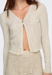 Femme portant un cardigan boutonné beige côtelé avec des bords festonnés et une jupe en dentelle florale beige.