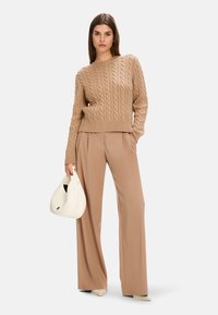 Beige Kabelstrickpullover mit Rundhalsausschnitt, kombiniert mit beigen weitem Hosen. Die Modells hält eine weiße Handtasche mit geschwungenem Design.