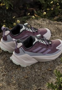 Paar lila und weiße Adidas Terrex Gore-Tex Trailrunning-Schuhe mit schwarzen Schnürsenkeln, die auf einer Kiesoberfläche im Freien liegen.