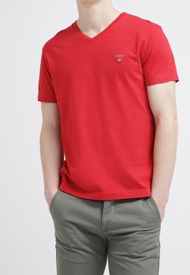 T-shirt en coton rouge avec col en V et manches courtes. Présente un petit logo GANT sur la poitrine. Porté avec un pantalon vert olive.