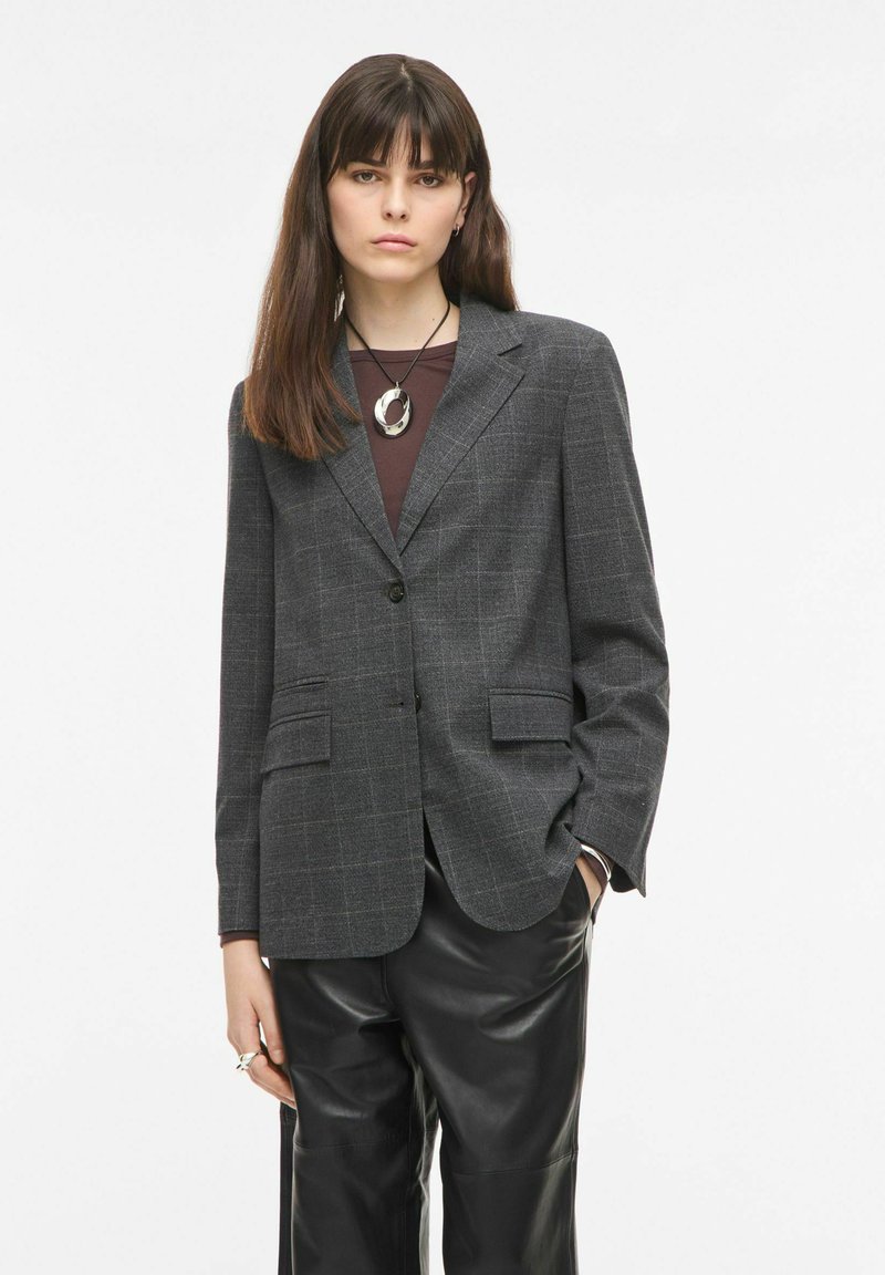 Jeune femme aux cheveux bruns et lisses portant un blazer gris à carreaux, un pantalon en cuir noir et un collier avec un pendentif circulaire.