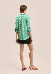 Chemise verte à motifs avec manches retroussées, associée à une jupe noire. Le mannequin porte des chaussures à talons ouvertes orange, mettant en valeur des lignes simples mais audacieuses.