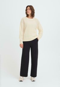 Kremfarget strikket genser med rund hals og teksturerte ermer, kombinert med svarte vide bukser og beige sneakers mot en ensfarget bakgrunn.