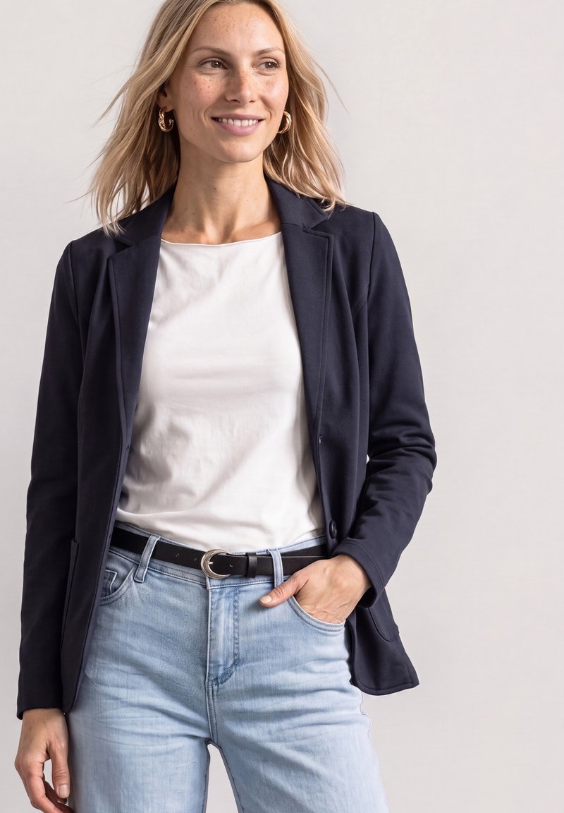 Femme souriante aux cheveux blonds, portant des boucles d'oreilles créoles en or, un blazer bleu marine sur un haut blanc, un jean bleu clair et une ceinture noire, main dans la poche.
