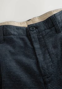 Ciemnoszare woskowane spodnie w kratę z zapięciem na guzik w pasie oraz beżową podszewką na jasnym tle.