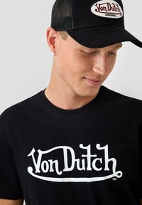 Juodas medvilnės marškinėliai su balta "Von Dutch" logotipu. Priedas - juoda tinklinė kepurė su balta ovalia patele ir raudonais užrašais.