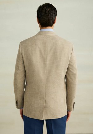 Muž otočený zády, oblečený v béžovém saku přes modrou košili a tmavě modré kalhoty, stojící před jednobarevným světlým pozadím.