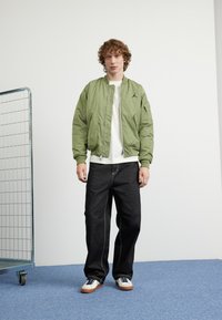 Giacca bomber verde chiaro con polsini a costine, logo nero sul petto, abbinata a jeans scuri e sneaker bianche su un pavimento blu.