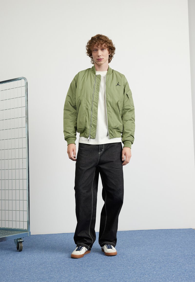 Chaqueta bomber verde claro con puños acanalados, logo negro en el pecho, combinada con jeans oscuros y zapatillas blancas sobre un suelo azul.