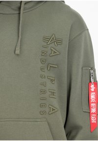 Sweat à capuche vert olive avec logo Alpha Industries en relief vertical sur la poitrine et étiquette rouge « Remove Before Flight » sur la poche zippée de la manche.