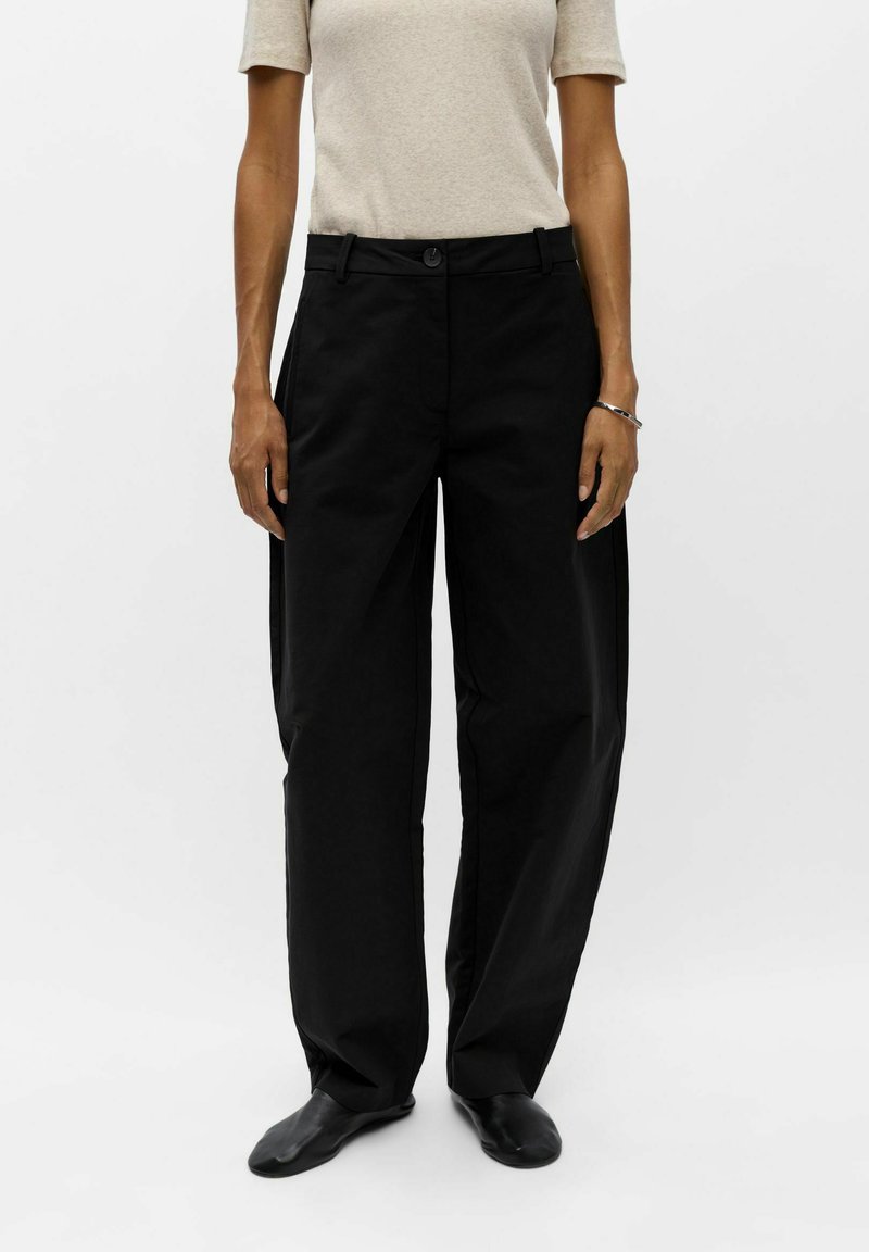 Personne portant un pantalon noir ample, un haut beige à manches courtes, des chaussures noires et un bracelet argenté, debout devant un fond blanc uni.
