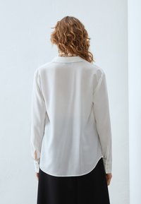 Weißes, langärmliges Blusenshirt mit Kragen, glatter Textur und seitlichen Schlitzen; kombiniert mit einem schwarzen Rock. Von hinten betrachtet.