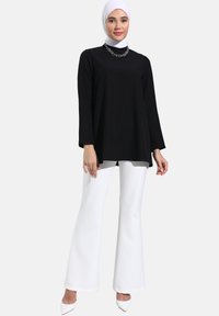 Tunic nera a maniche lunghe con scollatura rotonda, abbinata a pantaloni bianchi svasati e tacchi alti bianchi. Presenta una collana di catena argentata.