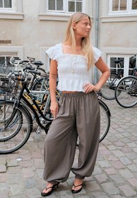Jeune femme aux longs cheveux blonds debout dans une rue pavée, portant un haut blanc à volants, un pantalon ample à carreaux et des sandales à talons noires près de vélos.