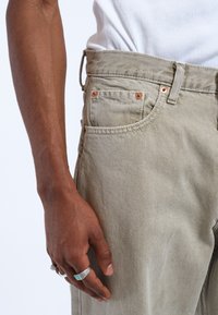 Helle khakifarbene Denim-Jeans mit kupferfarbenen Beschlägen, Fünf-Taschen-Design und strukturierter Oberfläche. Von Hand mit silbernen und türkisfarbenen Ringen verziert.