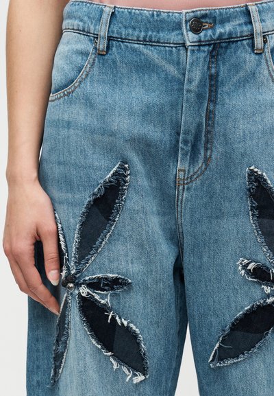 Jean en denim bleu clair à taille haute, avec de grandes découpes florales bordées de détails effilochés et des accents en denim foncé.
