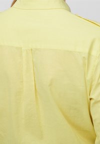 Camicia gialla leggera con colletto, dettaglio della piega posteriore e spalline, che mostra una texture liscia e una vestibilità rilassata.