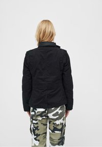 Giacca nera con un design strutturato e colletto alto. Indossata con pantaloni cargo mimetici che presentano diverse sfumature di verde e bianco.