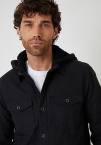 Czarna kurtka zapinana na guziki z kapturem, z miękkim kołnierzem, dwoma kieszeniami na piersi i teksturowaną podszewką. Noszona na białej koszulce.