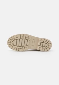 Suola beige di un stivale con profondi motivi di battistrada per grip, incluse scanalature ovali e circolari, posizionata su uno sfondo bianco uniforme.