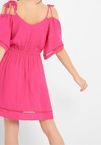 Vestido rosa con hombros al aire, mangas cortas, cintura elástica, con borde de encaje y tirantes atados en los hombros. Tejido ligero con una forma fluida.