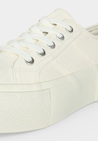 Gros plan d'une basket en toile blanche avec une semelle épaisse en caoutchouc et des lacets blancs passés dans des œillets métalliques.
