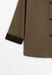 Giacca esterna marrone con una texture liscia, caratterizzata da una linea dritta, polsini neri a coste e una tasca laterale.