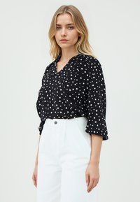 Blusa nera a pois con scollatura a V, maniche 3/4 e vestibilità comoda, realizzata in tessuto leggero. Indossata con pantaloni bianchi a vita alta.