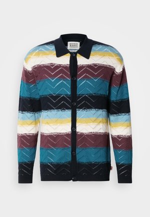 Pleteni kardigan s geometrijskim uzorkom naizmjeničnih pruga u tirkiznoj, burgundskoj, tamnoplavoj, žutoj i bijeloj boji, završen s kragnom i gumbima.