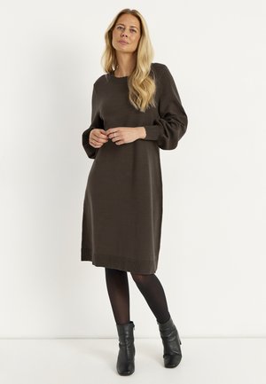 Kvinde med langt blondt hår iført en mørkebrun langærmet sweaterkjole, sorte strømpebukser og sorte ankelstøvler, stående mod en hvid baggrund.
