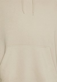 Beżowa bluza z kapturem wykonana z miękkiego materiału, z przednią kieszenią, ściągaczem w kapturze i widocznymi szwami na krawędziach.