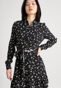Abito floreale nero con maniche lunghe, chiusura frontale con bottoni, orlo a ruffle e cintura a fiocco con il marchio "Tommy Jeans". Tessuto morbido e leggero.