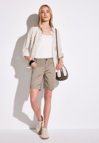 Chemise en lin beige par-dessus un débardeur blanc ; shorts kaki avec poches ; baskets marron et crème à motifs ; petit sac à bandoulière vert.