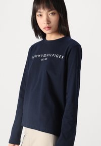 Sötétkék, hosszú ujjú póló, pamutból készült, középen fehér "TOMMY HILFIGER EST. 1985" logónyomattal. Egyszerű fazon, klasszikus dizájn.