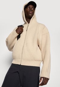 Felpa con zip color beige, a costine, con collo alto e cappuccio integrato. Caratterizzata da una vestibilità oversize e maniche lunghe. Zip nera e dettagli cuciti.
