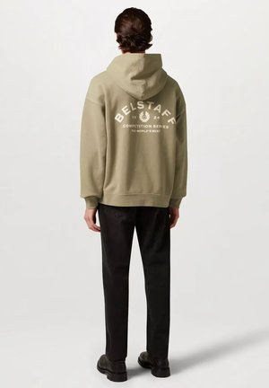 Persona con sudadera con capucha Belstaff de color verde oliva con la capucha puesta, pantalones negros y zapatos negros, vista desde atrás sobre un fondo liso.