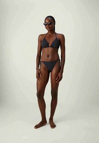 Mulher com pele escura e cabelo trançado, vestindo um biquíni preto e óculos de sol, de pé descalça contra um fundo claro liso.