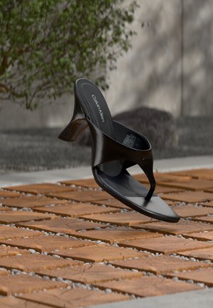 Sandale negre Calvin Klein cu toc și bareta în formă de șnur, plutind deasupra unei suprafețe exterioare acoperite cu dale, cu vegetație și un perete de beton în fundal.