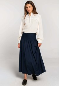 Blusa blanca de manga larga con tejido texturizado y volantes en la parte delantera, combinada con una falda larga de talle alto en color marino y mocasines negros.