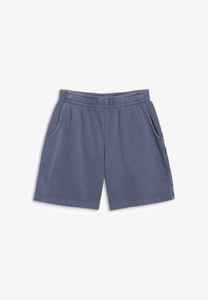 Fille souriante portant un sweat à capuche rose zippé, un t-shirt blanc, un short bleu, des chaussettes blanches avec des rayures noires et des baskets grises, debout sur un fond uni.