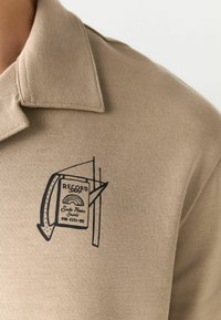Beige polo shirt featuring a black graphic design of a book labeled "RECORD" with a rainbow motif, and visible collar detail.